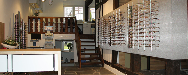 Heller Verkaufsraum eines Optikers mit großer Brillenauswahl an der Wand und Holztreppe.